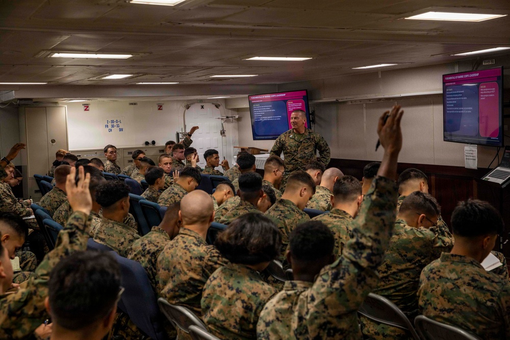 A military instructor in camouflage uniform conducts a classroom briefing for a large group of uniformed Marines in what appears to be a shipboard training room. Multiple attendees have their hands raised in response to a question or prompt from the instructor. Two display screens at the front show a presentation titled 'Fundamentals of Marine Corps Leadership'.