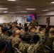 A military instructor in camouflage uniform conducts a classroom briefing for a large group of uniformed Marines in what appears to be a shipboard training room. Multiple attendees have their hands raised in response to a question or prompt from the instructor. Two display screens at the front show a presentation titled 'Fundamentals of Marine Corps Leadership'.
