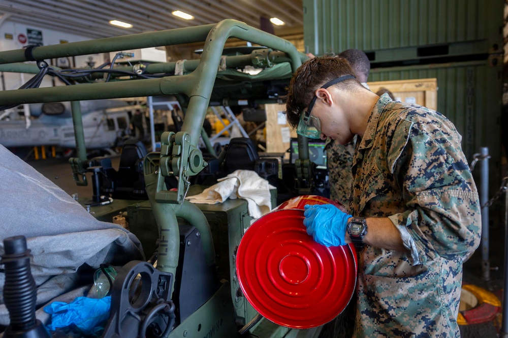 A U.S. Marine in digital camouflage (MARPAT) uniform is performing maintenance work on a military vehicle inside what appears to be a hangar or maintenance bay. The service member is wearing blue nitrile gloves and safety goggles while handling a bright red circular component, likely a fuel or oil container lid. Additional military equipment and what appears to be a helicopter are visible in the background.