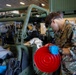 A U.S. Marine in digital camouflage (MARPAT) uniform is performing maintenance work on a military vehicle inside what appears to be a hangar or maintenance bay. The service member is wearing blue nitrile gloves and safety goggles while handling a bright red circular component, likely a fuel or oil container lid. Additional military equipment and what appears to be a helicopter are visible in the background.