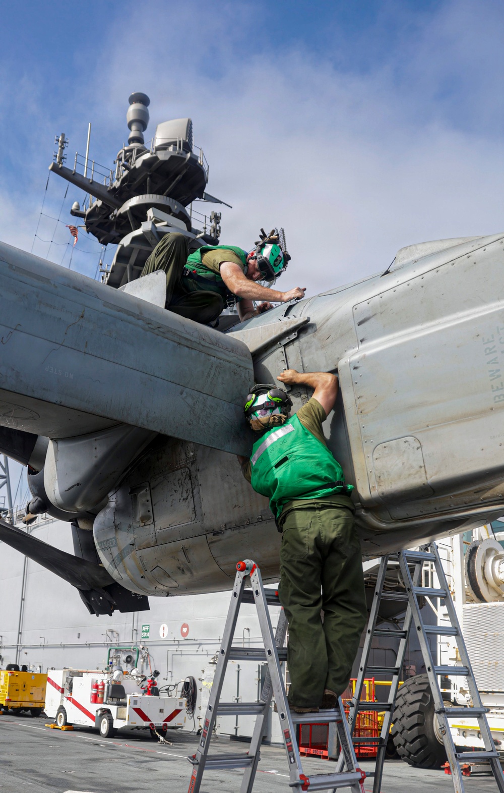 Two military maintenance personnel wearing green jerseys and helmets with hearing protection are performing maintenance work on a military aircraft on what appears to be the flight deck of a naval vessel. One technician is positioned on top of the aircraft while the other stands on a ladder below, both working on the same area of the fuselage or engine nacelle. The ship's island superstructure with radar equipment is visible in the background.
