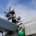 Two military maintenance personnel wearing green jerseys and helmets with hearing protection are performing maintenance work on a military aircraft on what appears to be the flight deck of a naval vessel. One technician is positioned on top of the aircraft while the other stands on a ladder below, both working on the same area of the fuselage or engine nacelle. The ship's island superstructure with radar equipment is visible in the background.