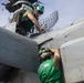 Two naval aviation personnel in green jerseys are performing maintenance on a military aircraft on what appears to be an aircraft carrier flight deck. One technician is positioned on top of the aircraft while the other works at a lower level near the fuselage. A radar array and carrier island superstructure are visible in the background.