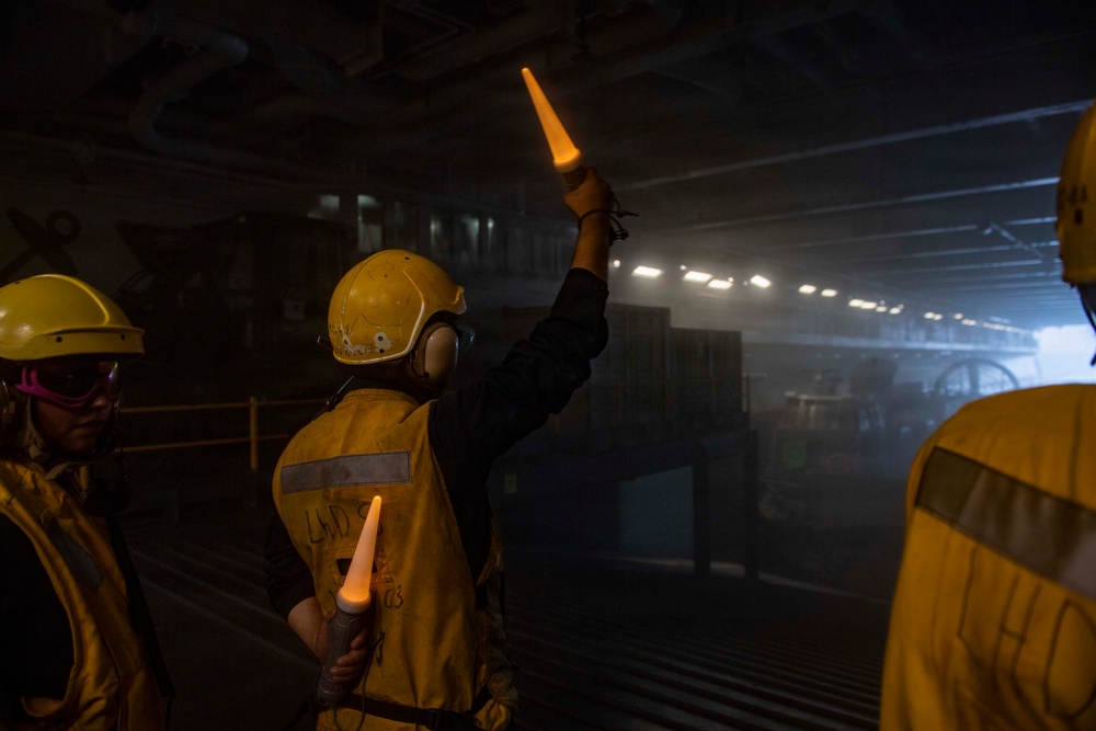 Personnel wearing yellow safety helmets, ear protection, and high-visibility vests are working in what appears to be a large enclosed hangar deck, likely aboard a naval vessel or aircraft carrier. The central figure is holding two illuminated orange wands/batons raised in a signaling gesture, consistent with aircraft or vehicle marshaling operations. The environment is dimly lit with hazy atmospheric conditions visible in the background.