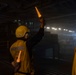 Personnel wearing yellow safety helmets, ear protection, and high-visibility vests are working in what appears to be a large enclosed hangar deck, likely aboard a naval vessel or aircraft carrier. The central figure is holding two illuminated orange wands/batons raised in a signaling gesture, consistent with aircraft or vehicle marshaling operations. The environment is dimly lit with hazy atmospheric conditions visible in the background.