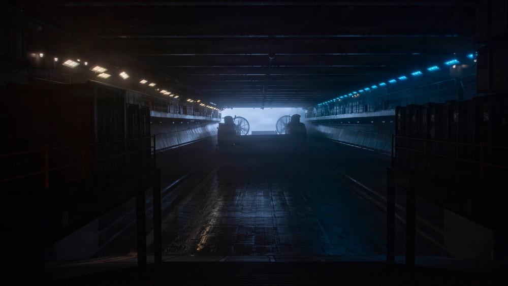 A dark, industrial interior space — likely the well deck of a naval amphibious assault ship — viewed from the stern looking toward an open rear gate with ambient exterior light visible. Two silhouetted craft, consistent with Landing Craft Air Cushion (LCAC) hovercraft, are positioned side by side near the opening. The scene is dramatically lit with rows of overhead lights on both sides and a blue-tinted light strip on the right wall.