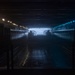 A dark, industrial interior space — likely the well deck of a naval amphibious assault ship — viewed from the stern looking toward an open rear gate with ambient exterior light visible. Two silhouetted craft, consistent with Landing Craft Air Cushion (LCAC) hovercraft, are positioned side by side near the opening. The scene is dramatically lit with rows of overhead lights on both sides and a blue-tinted light strip on the right wall.