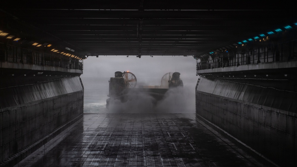 A Landing Craft Air Cushion (LCAC) hovercraft is entering or exiting the well deck of a naval amphibious assault ship. The image is taken from inside the well deck looking outward, with the hovercraft surrounded by spray and mist generated by its lift fans. The dramatic low-light composition highlights the industrial scale of the well deck structure.