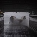 A Landing Craft Air Cushion (LCAC) hovercraft is entering or exiting the well deck of a naval amphibious assault ship. The image is taken from inside the well deck looking outward, with the hovercraft surrounded by spray and mist generated by its lift fans. The dramatic low-light composition highlights the industrial scale of the well deck structure.