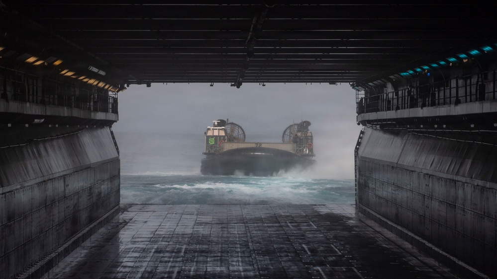 The image is taken from inside the well deck of a naval amphibious assault ship, looking outward toward an approaching Landing Craft Air Cushion (LCAC) hovercraft on rough, choppy seas. The LCAC is navigating through turbulent water and spray as it prepares to enter the ship's flooded well deck. The perspective emphasizes the massive scale of the well deck opening and the challenging sea conditions.