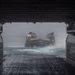 The image is taken from inside the well deck of a naval amphibious assault ship, looking outward toward an approaching Landing Craft Air Cushion (LCAC) hovercraft on rough, choppy seas. The LCAC is navigating through turbulent water and spray as it prepares to enter the ship's flooded well deck. The perspective emphasizes the massive scale of the well deck opening and the challenging sea conditions.