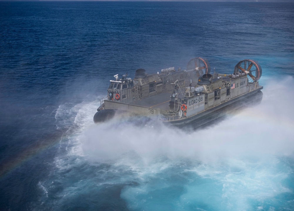 The image shows a U.S. Navy Landing Craft Air Cushion (LCAC-39) operating at high speed over open ocean water. The hovercraft is generating a large spray of white mist and foam beneath and around its hull as it skims across the surface. The photograph is taken from an elevated aerial perspective, clearly showing the vessel's designation markings and equipment.