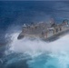 The image shows a U.S. Navy Landing Craft Air Cushion (LCAC-39) operating at high speed over open ocean water. The hovercraft is generating a large spray of white mist and foam beneath and around its hull as it skims across the surface. The photograph is taken from an elevated aerial perspective, clearly showing the vessel's designation markings and equipment.
