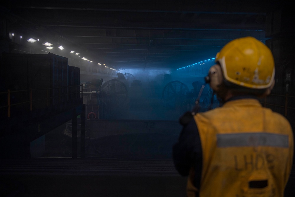 A worker wearing a yellow hard hat, hearing protection, and a high-visibility vest marked 'LHD8' observes a large, dimly lit interior space — likely the well deck or vehicle deck of a naval amphibious assault ship (LHD-8, USS Makin Island). The space is filled with haze or smoke and illuminated by rows of overhead lights casting a blue tint. Large mechanical equipment, possibly vehicles or machinery with visible wheels, can be seen in the background.