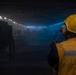 A worker wearing a yellow hard hat, hearing protection, and a high-visibility vest marked 'LHD8' observes a large, dimly lit interior space — likely the well deck or vehicle deck of a naval amphibious assault ship (LHD-8, USS Makin Island). The space is filled with haze or smoke and illuminated by rows of overhead lights casting a blue tint. Large mechanical equipment, possibly vehicles or machinery with visible wheels, can be seen in the background.