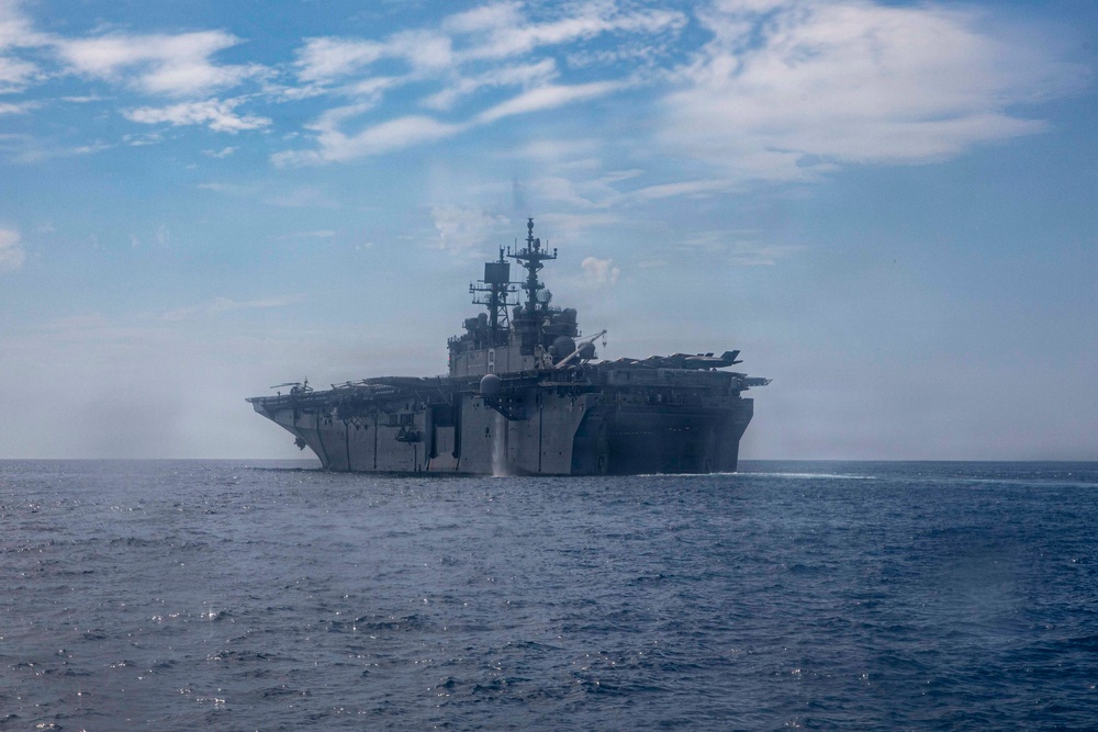 A large US Navy amphibious assault ship is photographed at sea from a bow-on perspective, showing its flat flight deck and island superstructure. The vessel appears to be underway in open ocean under partly cloudy skies. Steam or exhaust is visible rising from the ship's superstructure area.