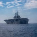 A large US Navy amphibious assault ship is photographed at sea from a bow-on perspective, showing its flat flight deck and island superstructure. The vessel appears to be underway in open ocean under partly cloudy skies. Steam or exhaust is visible rising from the ship's superstructure area.