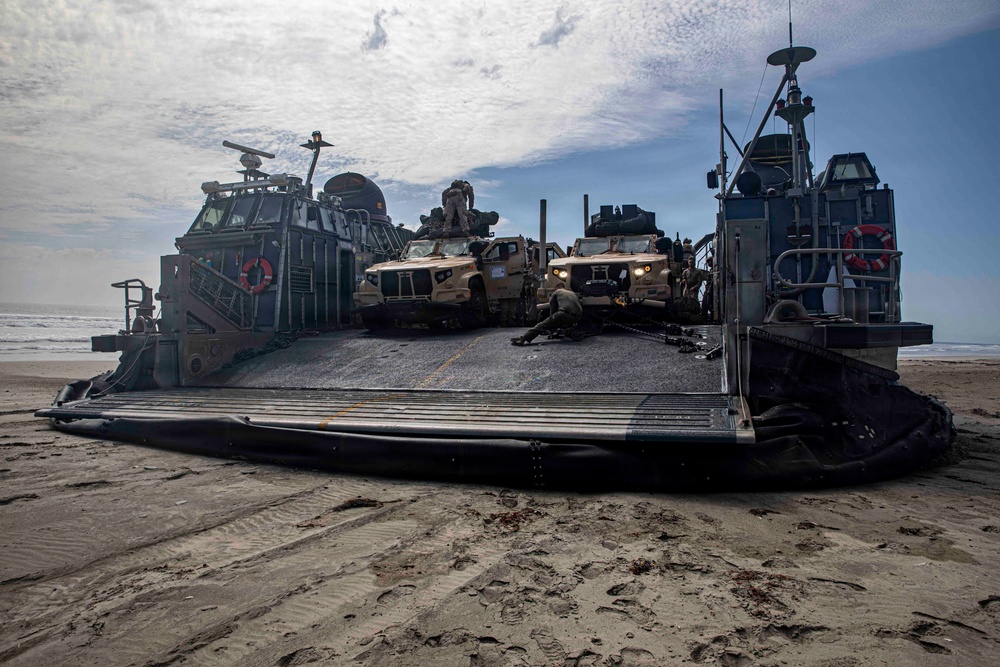 A military Landing Craft Air Cushion (LCAC) hovercraft has beached on a sandy shoreline with its bow ramp lowered. The craft is loaded with multiple tan/coyote-colored military tactical vehicles, likely Joint Light Tactical Vehicles (JLTVs), with personnel visible on and around them. This appears to be an amphibious assault or beach landing exercise.