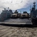 A military Landing Craft Air Cushion (LCAC) hovercraft has beached on a sandy shoreline with its bow ramp lowered. The craft is loaded with multiple tan/coyote-colored military tactical vehicles, likely Joint Light Tactical Vehicles (JLTVs), with personnel visible on and around them. This appears to be an amphibious assault or beach landing exercise.