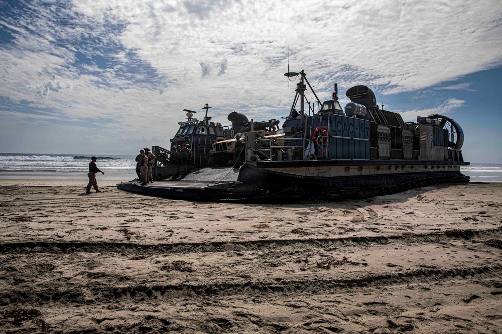 A U.S. Navy Landing Craft Air Cushion (LCAC) designated LCAC-167 is beached on a sandy shoreline, likely during an amphibious landing operation or exercise. Several personnel in military uniforms are visible near the craft's ramp, and at least one military vehicle is loaded on the deck. The image is taken in strong backlit conditions, creating silhouette effects against a partly cloudy sky.
