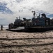 A U.S. Navy Landing Craft Air Cushion (LCAC) designated LCAC-167 is beached on a sandy shoreline, likely during an amphibious landing operation or exercise. Several personnel in military uniforms are visible near the craft's ramp, and at least one military vehicle is loaded on the deck. The image is taken in strong backlit conditions, creating silhouette effects against a partly cloudy sky.