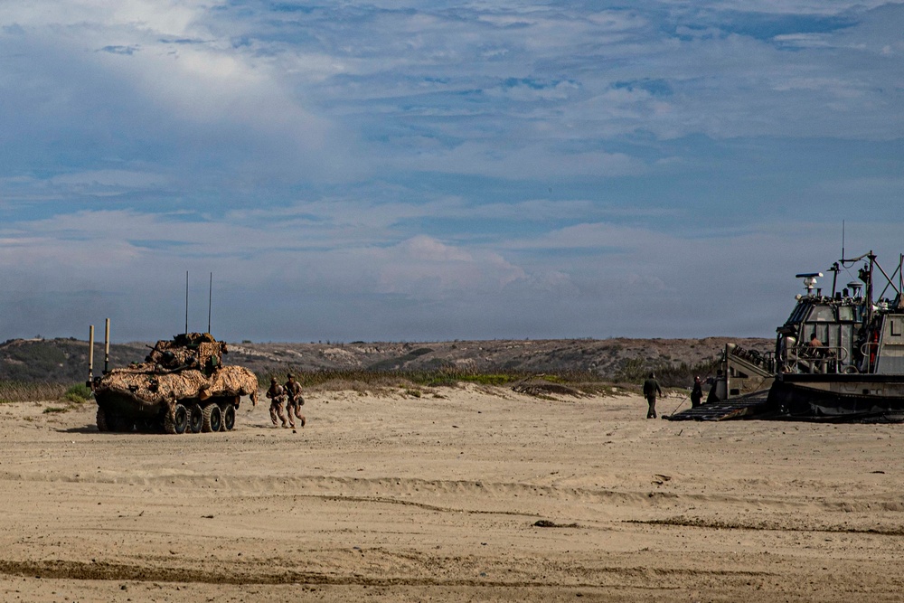A military amphibious operation is underway on a sandy beach, featuring a camouflaged wheeled armored vehicle (likely an LAV-25 or similar) on the left and a landing craft on the right. Several personnel in military uniforms are visible between the two vehicles on the beach. The terrain consists of sandy dunes with sparse vegetation under a partly cloudy sky.