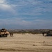 A military amphibious operation is underway on a sandy beach, featuring a camouflaged wheeled armored vehicle (likely an LAV-25 or similar) on the left and a landing craft on the right. Several personnel in military uniforms are visible between the two vehicles on the beach. The terrain consists of sandy dunes with sparse vegetation under a partly cloudy sky.