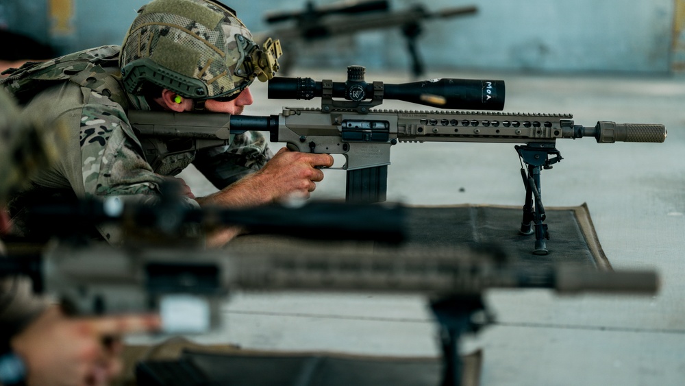 Military personnel are engaged in a precision rifle shooting exercise at what appears to be an indoor or covered firing range. The primary subject is a soldier in full combat gear aiming a designated marksman rifle equipped with a high-powered scope, bipod, and suppressor. At least two shooters are visible, suggesting a coordinated marksmanship training session.