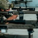 Military personnel are engaged in a precision rifle shooting exercise at what appears to be an indoor or covered firing range. The primary subject is a soldier in full combat gear aiming a designated marksman rifle equipped with a high-powered scope, bipod, and suppressor. At least two shooters are visible, suggesting a coordinated marksmanship training session.