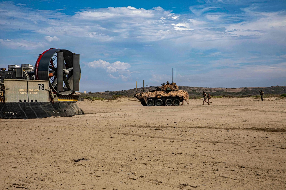 A military beach landing operation is depicted, showing a Landing Craft Air Cushion (LCAC) hovercraft numbered '78' on the left and a wheeled armored fighting vehicle in desert camouflage in the center-background. Several military personnel are visible near the armored vehicle on a sandy beach terrain. The scene appears to be an amphibious assault exercise or operation.