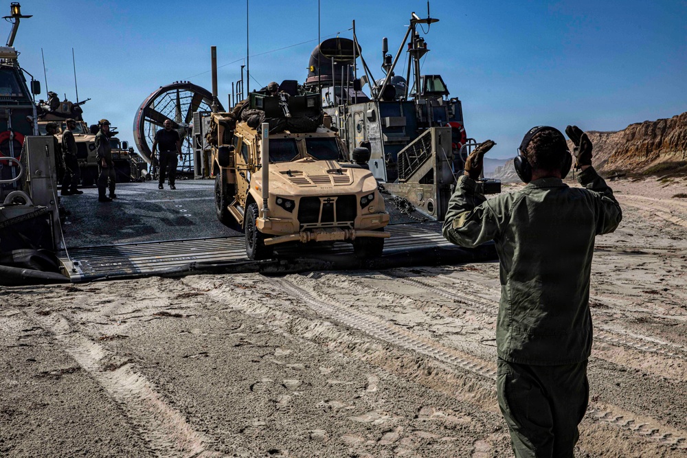 A military amphibious landing craft (LCAC - Landing Craft Air Cushion) has beached on a sandy shoreline, and a tan/desert-colored MRAP-type tactical vehicle (appears to be a Joint Light Tactical Vehicle or similar) is being offloaded down the ramp. A military personnel member in olive green flight suit and hearing protection is directing the vehicle with hand signals. The terrain features sandy beach with rocky cliffs visible in the background under clear blue skies.
