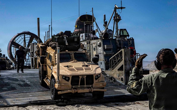 A military amphibious landing craft (LCAC - Landing Craft Air Cushion) has beached on a sandy shoreline, and a tan/desert-colored MRAP-type tactical vehicle (appears to be a Joint Light Tactical Vehicle or similar) is being offloaded down the ramp. A military personnel member in olive green flight suit and hearing protection is directing the vehicle with hand signals. The terrain features sandy beach with rocky cliffs visible in the background under clear blue skies.