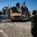 A military amphibious landing craft (LCAC - Landing Craft Air Cushion) has beached on a sandy shoreline, and a tan/desert-colored MRAP-type tactical vehicle (appears to be a Joint Light Tactical Vehicle or similar) is being offloaded down the ramp. A military personnel member in olive green flight suit and hearing protection is directing the vehicle with hand signals. The terrain features sandy beach with rocky cliffs visible in the background under clear blue skies.