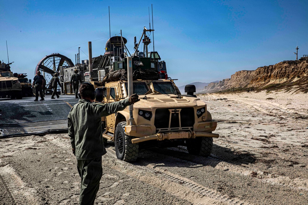 A military beach landing operation is underway, with a tan/desert-colored Joint Light Tactical Vehicle (JLTV) being guided off what appears to be a Navy Landing Craft Air Cushion (LCAC) hovercraft onto a sandy beach. A service member in olive drab flight suit is directing the vehicle with hand signals. The terrain features sandy beach with rocky cliffs visible in the background under clear blue skies.