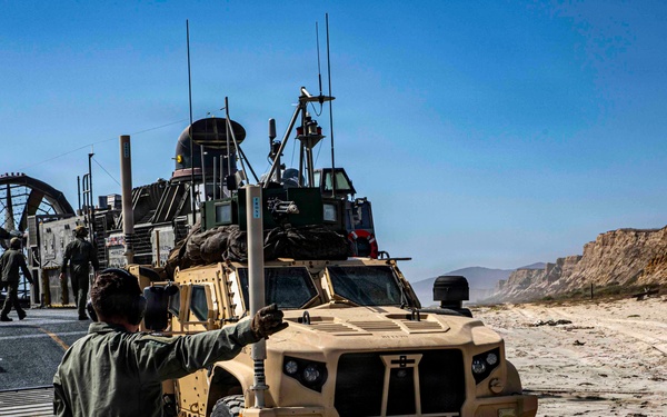 A military beach landing operation is underway, with a tan/desert-colored Joint Light Tactical Vehicle (JLTV) being guided off what appears to be a Navy Landing Craft Air Cushion (LCAC) hovercraft onto a sandy beach. A service member in olive drab flight suit is directing the vehicle with hand signals. The terrain features sandy beach with rocky cliffs visible in the background under clear blue skies.