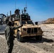 A military beach landing operation is underway, with a tan/desert-colored Joint Light Tactical Vehicle (JLTV) being guided off what appears to be a Navy Landing Craft Air Cushion (LCAC) hovercraft onto a sandy beach. A service member in olive drab flight suit is directing the vehicle with hand signals. The terrain features sandy beach with rocky cliffs visible in the background under clear blue skies.