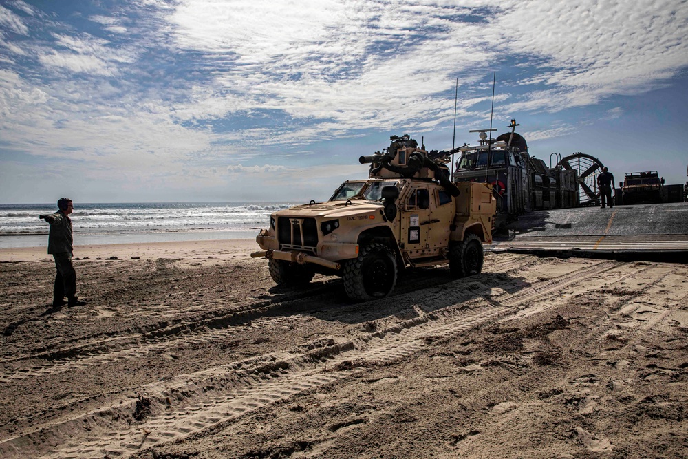 A military amphibious landing operation is underway on a sandy beach, with a tan/desert-colored Joint Light Tactical Vehicle (JLTV) having disembarked from a Landing Craft Air Cushion (LCAC) hovercraft visible in the background. A service member on the left appears to be directing vehicle traffic. The scene shows tire tracks in the sand and additional military vehicles still on or near the landing craft.