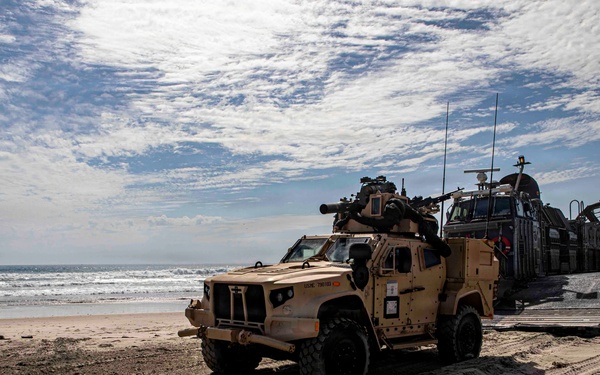 A military amphibious landing operation is underway on a sandy beach, with a tan/desert-colored Joint Light Tactical Vehicle (JLTV) having disembarked from a Landing Craft Air Cushion (LCAC) hovercraft visible in the background. A service member on the left appears to be directing vehicle traffic. The scene shows tire tracks in the sand and additional military vehicles still on or near the landing craft.