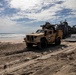 A military amphibious landing operation is underway on a sandy beach, with a tan/desert-colored Joint Light Tactical Vehicle (JLTV) having disembarked from a Landing Craft Air Cushion (LCAC) hovercraft visible in the background. A service member on the left appears to be directing vehicle traffic. The scene shows tire tracks in the sand and additional military vehicles still on or near the landing craft.