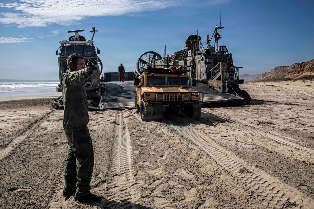 A military beach landing operation is underway, with a Landing Craft Air Cushion (LCAC) hovercraft beached on a sandy shoreline. A tan/desert-colored HMMWV (Humvee) is being offloaded from the craft's ramp onto the beach. A military personnel member in olive drab uniform is directing the vehicle's movement using hand signals.