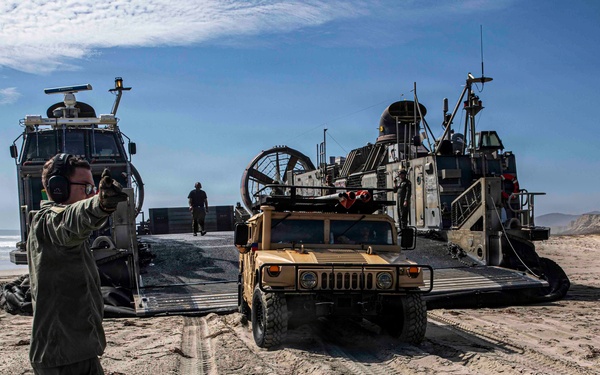 A military beach landing operation is underway, with a Landing Craft Air Cushion (LCAC) hovercraft beached on a sandy shoreline. A tan/desert-colored HMMWV (Humvee) is being offloaded from the craft's ramp onto the beach. A military personnel member in olive drab uniform is directing the vehicle's movement using hand signals.