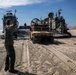 A military beach landing operation is underway, with a Landing Craft Air Cushion (LCAC) hovercraft beached on a sandy shoreline. A tan/desert-colored HMMWV (Humvee) is being offloaded from the craft's ramp onto the beach. A military personnel member in olive drab uniform is directing the vehicle's movement using hand signals.