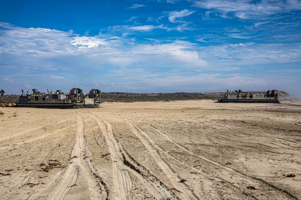 Two U.S. Navy Landing Craft Air Cushion (LCAC) hovercraft are beached on a sandy shoreline, likely during an amphibious landing exercise or operation. The vehicles show distinctive large propeller fans with red-accented shrouds and military markings. Heavy tire tracks in the sand indicate recent vehicle movement from the landing craft.