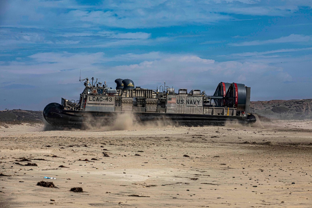A U.S. Navy Landing Craft Air Cushion (LCAC-43) hovercraft is traversing a sandy beach, kicking up dust and sand beneath its air cushion skirt. The vessel bears clear U.S. Navy markings and an American flag on its hull. The scene appears to be a military amphibious landing exercise or operational beach approach.