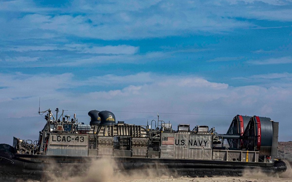 A U.S. Navy Landing Craft Air Cushion (LCAC-43) hovercraft is traversing a sandy beach, kicking up dust and sand beneath its air cushion skirt. The vessel bears clear U.S. Navy markings and an American flag on its hull. The scene appears to be a military amphibious landing exercise or operational beach approach.