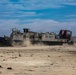 A U.S. Navy Landing Craft Air Cushion (LCAC-43) hovercraft is traversing a sandy beach, kicking up dust and sand beneath its air cushion skirt. The vessel bears clear U.S. Navy markings and an American flag on its hull. The scene appears to be a military amphibious landing exercise or operational beach approach.