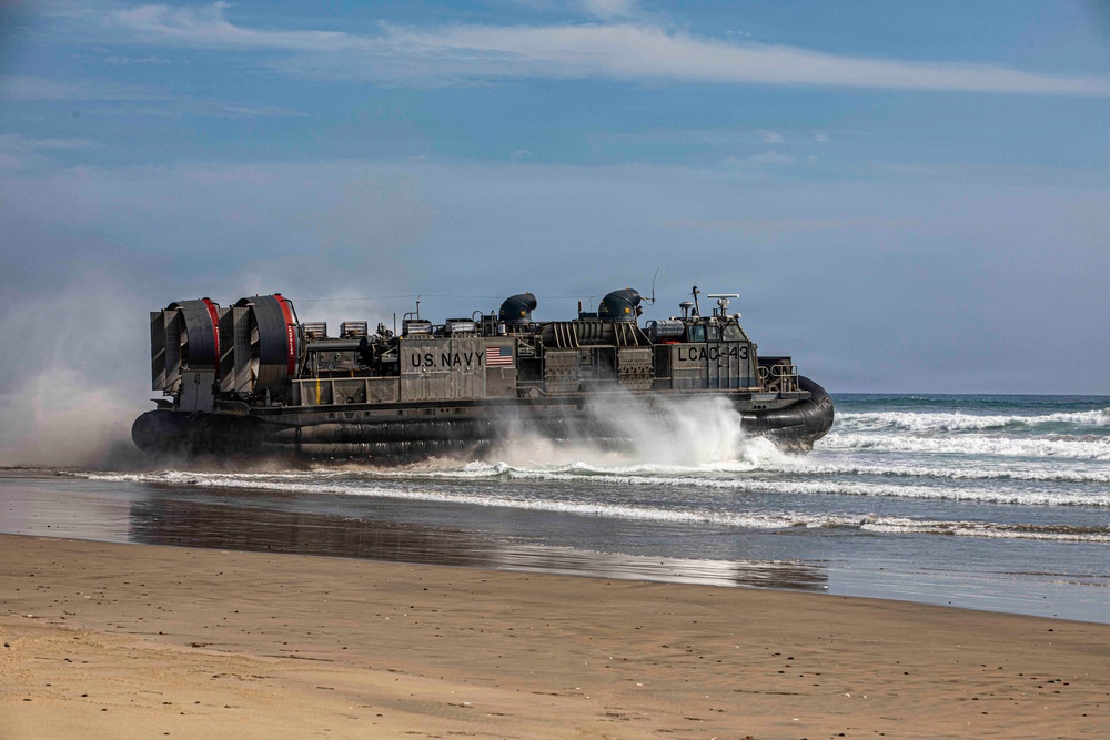 A U.S. Navy Landing Craft Air Cushion (LCAC) designated LCAC-43 is coming ashore on a sandy beach, transitioning from water to land. The hovercraft is generating significant spray and mist as it crosses the surf zone. This appears to be an amphibious landing operation or training exercise.