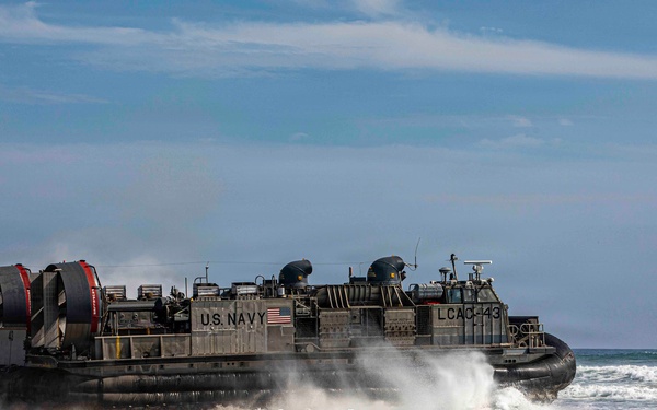 A U.S. Navy Landing Craft Air Cushion (LCAC) designated LCAC-43 is coming ashore on a sandy beach, transitioning from water to land. The hovercraft is generating significant spray and mist as it crosses the surf zone. This appears to be an amphibious landing operation or training exercise.