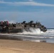 A U.S. Navy Landing Craft Air Cushion (LCAC) designated LCAC-43 is coming ashore on a sandy beach, transitioning from water to land. The hovercraft is generating significant spray and mist as it crosses the surf zone. This appears to be an amphibious landing operation or training exercise.