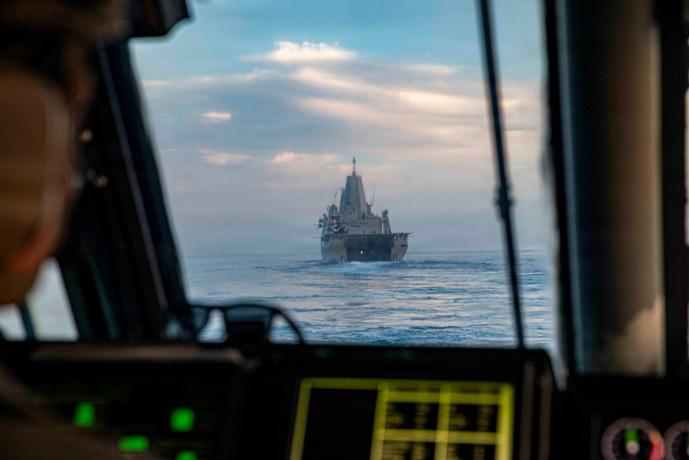 The image is taken from the cockpit of a military aircraft or helicopter, looking through the windscreen at a large naval vessel underway at sea. A person in the foreground, likely a pilot or crew member, is partially visible on the left side. The naval vessel appears to be a US Navy amphibious transport dock (LPD-class) ship, viewed from the bow/front aspect.